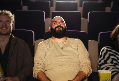 Charlie sits in a cinema between two people. He is looking up at the screen and smiling. He has a shaved head and a dark brown beard.