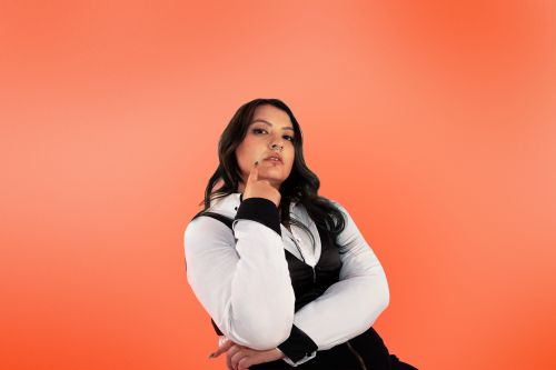 Gabby stands in a
photography studio posing for
the camera. She stands in front
of an orange backdrop
wearing a white shirt with a
black vest.
