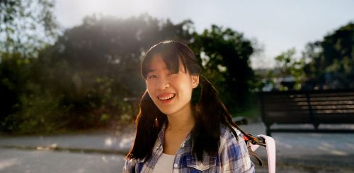 Nicole sits in a wheelchair outdoors on a sunny day, slightly turned and smiling at the camera. A bench and greenery are blurred in the background. She wears a blue and white plaid shirt over a white T-shirt and has dark brown hair in two pigtails.