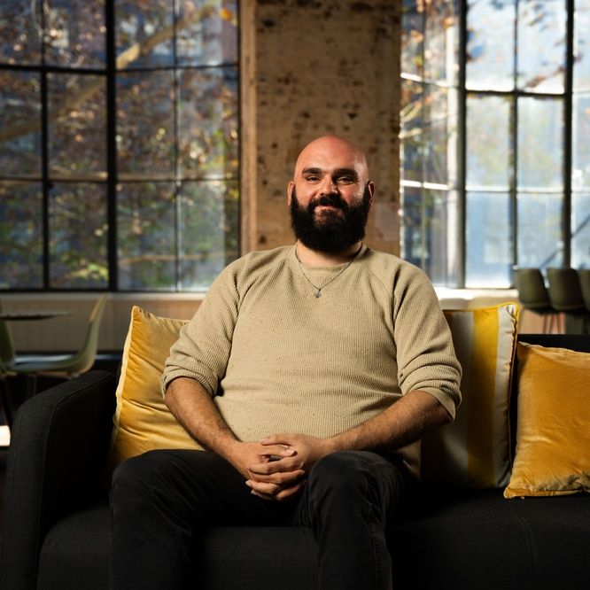 Charlie sits on a couch in an open plan living area smiling at the camera while wearing a cream-coloured jumper. He has a shaved head and a dark brown beard.