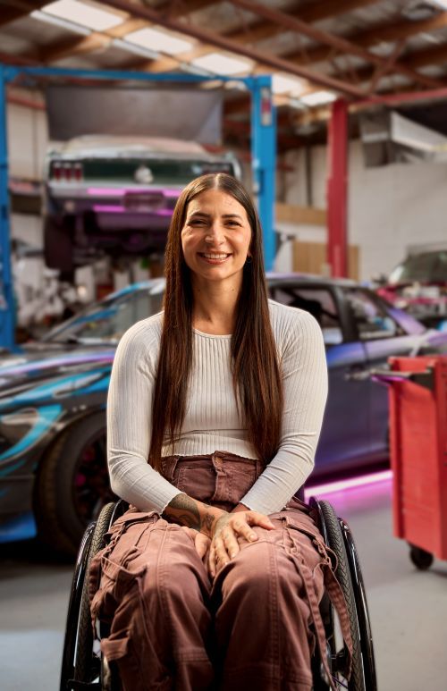 Christina smiles at the camera with her long straight brown hair down. She is wearing a white long-sleeved top and her purple drift car is parked behind her in a mechanic workshop.