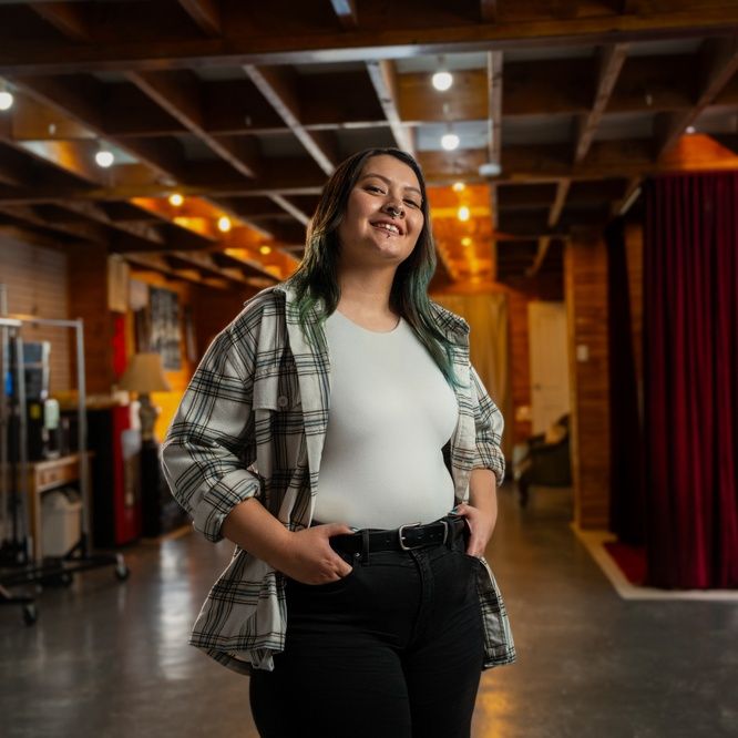 Gabby stands in a photography studio smiling at the camera, with her hands in the pockets of her jeans. A red curtain and photography equipment are visible behind her.