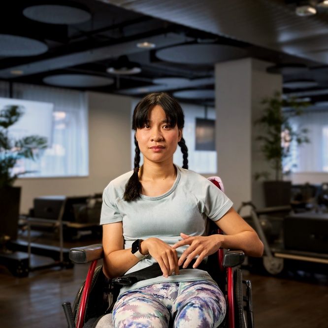 Nicole sits in a wheelchair in a Pilates studio, wearing a light blue T-shirt and smiling slightly at the camera. She has dark brown hair styled in two braids.