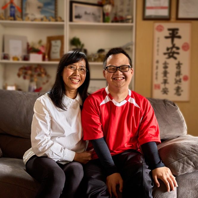 Nino and his mother Kim sit together on a grey couch in a lounge room, smiling toward the camera with shelves and various home decor behind them.