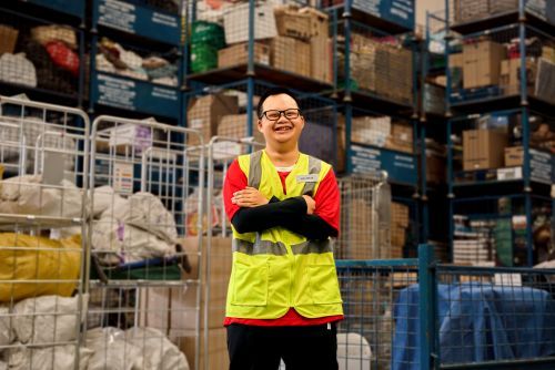 Nino stands in a warehouse wearing glasses and a hi-vis work shirt, smiling with his arms crossed.