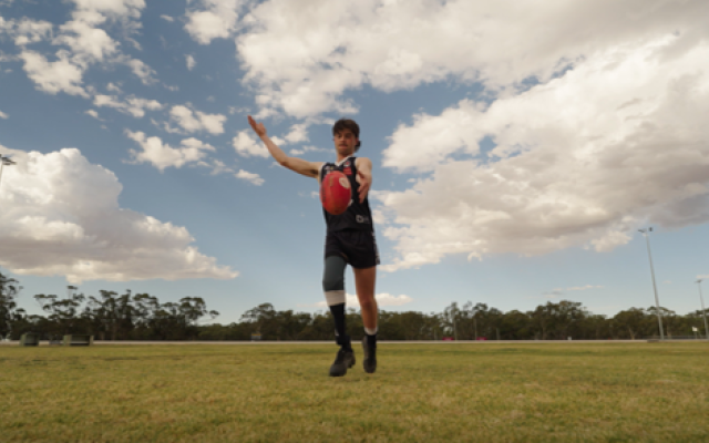 Person in sports uniform holding a red Australian rules football while moving across a grassy sports field under a partly cloudy sky.