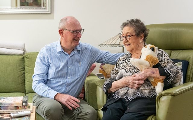 A person sitting beside an older person in a lounge room, talking while the older person holds a soft toy in an armchair.