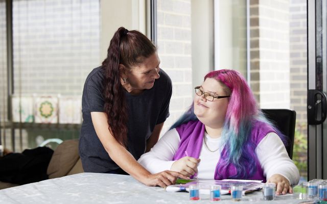 image of a person sitting and painting and another standing and showing them something on a page
